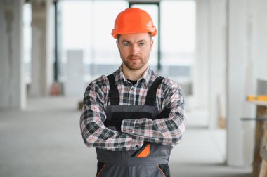 Turuncu kask takan kendine güvenen bir inşaatçının portresi ve inşaat halindeki bir binada çapraz kollarla duran iş tulumları.