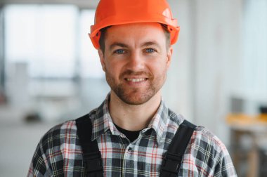 İnşaat işçisi turuncu bir kask takıyor ve inşaat halindeki bir binanın içindeki kameraya gülümsüyor.