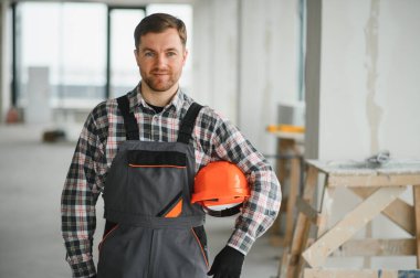 İnşaat işçisi miğferini tutuyor ve inşaat halindeki bir binada çalışmaya hazırlanırken iş tulumu giyiyor.