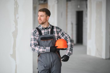 Üniformalı bir inşaat işçisi kolunun altında turuncu bir miğfer tutuyor ve inşaat alanındaki gelişmeleri inceliyor.