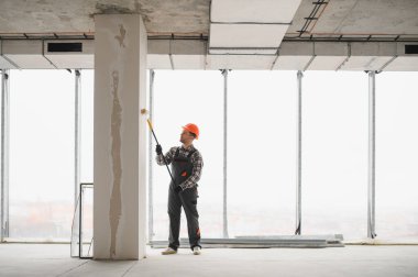 İnşaat işçisi koruyucu kask ve tulum giyiyor, uzun bir silindir kullanarak duvarı boyuyor, tadilat sırasında...