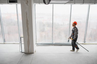 İnşaat işçisi inşaat halindeki bir binada yürüyor, aletler taşıyor ve güvenlik kaskı takıyor.