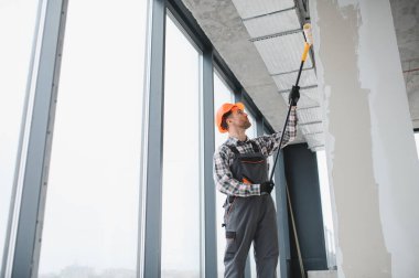 İnşaat işçisi inşaat halindeki bir binanın duvarlarını ve tavanını boyamak için uzun bir boya silindiri kullanıyor.