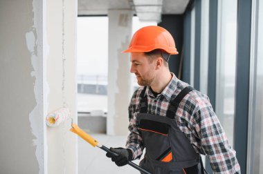 Modern bir binanın içindeki duvarları silindirle boyamak için koruyucu ekipman kullanan inşaatçı yenileme ve inşaat işlerine odaklanıyor.