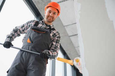Gülen müteahhit tekerlekli sandalye ve teleskopik saplı bir duvar boyuyor, iş tulumu ve güvenlik kaskı takıyor.