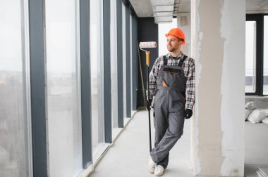 Boyacı tulum giyiyor ve şapka takıyor. Duvara yaslanırken modern bina yapım aşamasındaydı.