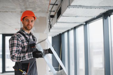 İnşaatçı güvenlik kaskı takıyor, inşaat halindeki bir binaya havalandırma sistemi kuruyor, uygun hava akışını ve konforu sağlıyor.