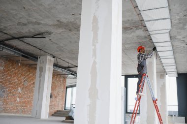 Güvenlik kaskı takan inşaat işçisi, merdivenin üstünde duruyor ve inşaat halindeki bir binaya havalandırma sistemi kuruyor.