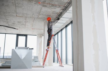 İnşaat işçisi inşaat halindeki bir binaya havalandırma döşeyen, güvenlik kaskı takan ve iş tulumu giyen bir merdivende çalışıyor.