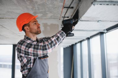 İnşaat işçisi güvenlik kaskı takıyor ve inşaat halindeki binaya havalandırma sistemi kurmak için matkap kullanıyor.