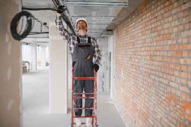 Bir inşaat alanında havalandırma ve havalandırma sistemi üzerinde çalışan elektrikçi, güvenlik kaskı takıyor ve genel olarak