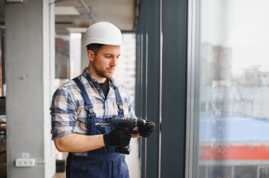 Güvenlik kaskı ve eldiven takan inşaat işçisi modern binanın penceresinin yakınına kablosuz matkap takıyor.