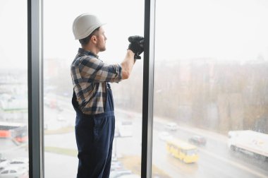Güvenlik kaskı takan ve kablosuz tornavida kullanan inşaat işçisi, modern bir binaya pencere takıyor.