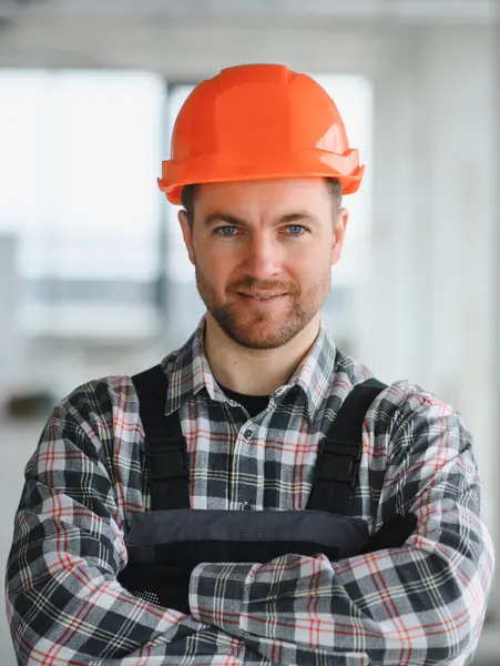 Turuncu sapkalı ve tulumlu kendine güvenen inşaat işçisi inşaat halindeki bir binada çapraz kollarla duruyor.