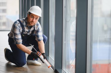 Koruyucu kask ve eldiven takan inşaat işçisi, yalıtım ve hava geçirmez olarak silikon dolgu malzemesi kullanarak pencere çerçevesini mühürlüyor.