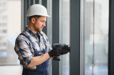 İnşaat işçisi, güvenlik kaskı takıyor ve iş tulumu giyiyor. İnşaat halindeki bir binaya yeni bir pencere takmak için kablosuz matkap kullanıyor.