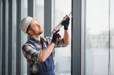 Güvenlik kaskı ve eldiven takan inşaat işçisi inşaat halindeki bir binanın pencere çerçevesine silikon dolgu maddesi sürüyor.