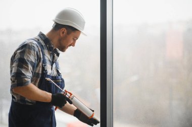 İnşaat işçisi koruyucu kask ve eldiven takıyor ve bina tadilatı sırasında pencere çerçevesine dolgu macunu sürüyor.