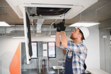 Güvenlik kaskı takan ve işçi tulumu giyen Hvac teknisyeni, modern ofiste elektrikli matkap kullanıyor, havalandırma sistemini tamir ediyor.