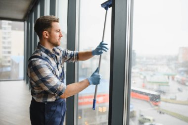 Mavi eldiven giyen temizlikçi modern bir ofis binasında camları teleskopik cam sileceğiyle yıkıyor.