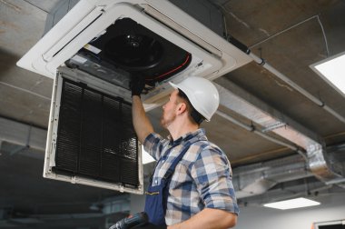 Kasket takan ve elinde tornavida tutan HVC teknisyeni tavandaki kaset tipi klima ünitesini tamir ediyor.