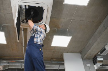 Güvenlik kaskı takan HVC teknisyeni ve havalandırmayı tamir eden üniformalı biri.