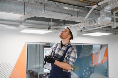 Güvenlik kaskı ve eldiven takan HVC teknisyeni, modern ofis binasındaki bakım ve tesisat sırasında havalandırma sistemini inceliyor.