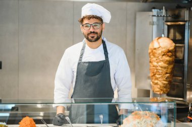 Fast food restoranının mutfağında, üniformalı bir ortadoğulu şef ustalıkla shawarma ve kebap hazırlıyor.