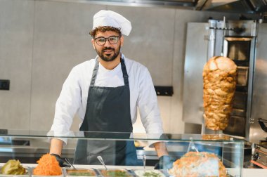 Ortadoğulu şef üniforma ve eldiven giyiyor, cam tezgahın arkasında çeşitli malzemelerle duruyor, modern bir restoran mutfağında shawarma hazırlıyor.