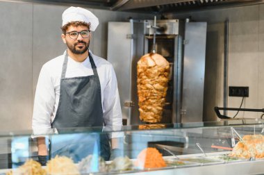 Profesyonel aşçı önlüğü ve toka giyip modern bir mutfakta leziz sokak yemekleri sunmaya hazır bir şavurma ya da kebap hazırlıyor.