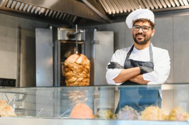 Kendine güvenen şef, kebap lokantasında çapraz kollarla poz veriyor. Lezzetli bir shawarma sergiliyor. Dikey bir rosto ve taze malzemeler üzerinde dönüyor.