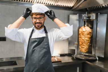 Gülümseyen şef şapkasını düzeltiyor ve ticari bir mutfakta siyah eldivenler giyiyor. Arka planda da da shawarma kızartması var.