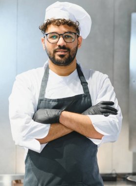 Kolları kavuşturulmuş siyah eldivenler giyen gülümseyen bir şefin portresi modern bir mutfakta aşçılık sanatına olan tutkusunu ve profesyonelliğini gösteriyor.