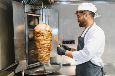 Profesyonel bir şef, ticari bir mutfakta lezzetli shawarma etini dönüşümlü bir tükürükten ustalıkla dilimliyor. Popüler bir sokak yemeği hazırlıyor.