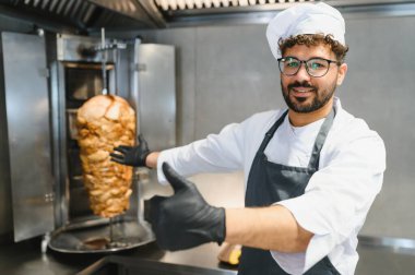 Gülümseyen şef leziz bir şavurma sunar. Dikey bir rostoda döner, baş parmağını kaldırır, siyah eldiven ve önlük giyer.