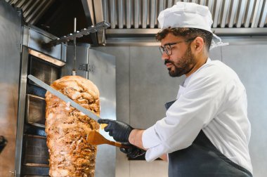 Profesyonel bir şef, ticari bir mutfakta lezzetli shawarma etini dönüşümlü bir tükürükten ustalıkla dilimliyor. Popüler bir orta doğu sokak yemeği hazırlıyor.