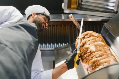 Profesyonel bir şef, ticari bir mutfakta döner tükürükten shawarma etini ustalıkla dilimliyor. Popüler bir sokak yemeği hazırlıyor.