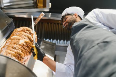Lezzetli shawarma pişirmek, dikey tükürük üzerinde et dilimlemek, ticari mutfakta aşçı şapkası ve önlüğü giymek.