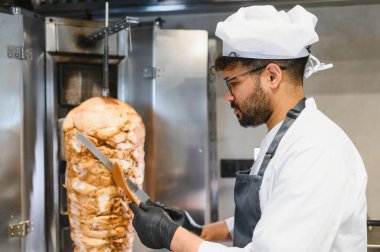 Profesyonel bir şef lezzetli shawarma etini dönüşümlü tükürükten ustalıkla dilimliyor, kalabalık bir restoranın mutfağında popüler bir sokak yemeği hazırlıyor.