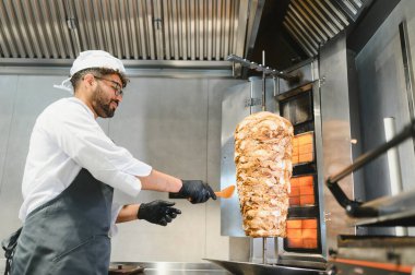 Aşçı, spatula kullanarak profesyonel bir mutfakta dikey bir rostoda dönen shawarma etinin yapısını kontrol eder.