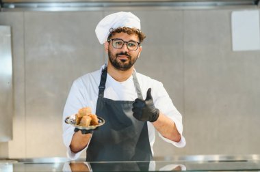 Kendine güvenen aşçı lezzetli baklavaları gösteriyor ve parmaklarını işaret ediyor. Profesyonel mutfakta siyah eldiven ve önlük giyiyor.