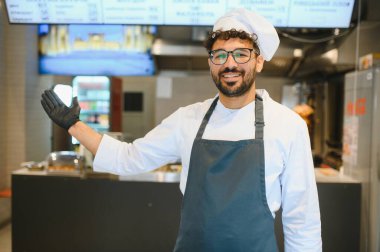 Dost canlısı şef, kebap restoranına gelen müşterileri karşılıyor. Üniformalı ve siyah eldivenli, tezgaha doğru hareket ediyor.