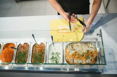 Aşçı pide ekmeğe et koymak için maşa kullanıyor, ön planda soslar ve sebzeler var.