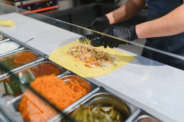 Tavuk ve patates kızartması ile bir shawarma 'ya havuç ve lahana ekleyen aşçı mutfak maşası kullanarak, metal kaplar dolusu sos ve malzemelerle dolu cam bir camın arkasında.