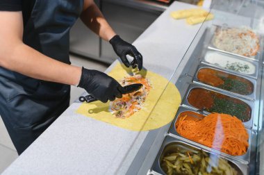 Aşçı pide ekmeğe sebze ve et ekler, maşa kullanır, shawarma ya da kebap hazırlar, arka planda malzemelerle dolu kaplar hazırlar.