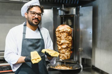 Siyah eldiven ve önlük takan aşçı, bir restoranın mutfağında iki shawarma tutuyor.