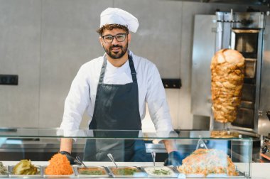 Orta Doğulu aşçı bir elbise ve önlük giyerek ticari bir mutfakta shawarma ve diğer yemekleri hazırlıyor.