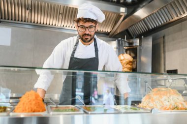 Üniforma ve siyah eldiven giyen genç aşçı cam tezgahın arkasında durup shawarma ya da kebap gibi Arap sokak yemekleri hazırlıyor.