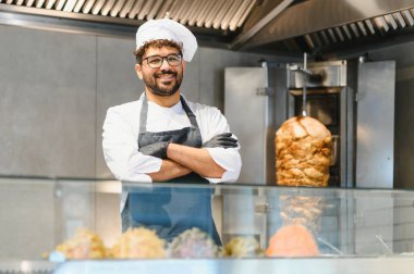 Kendine güvenen şef sokak yemekleri işinde çapraz kollarla poz veriyor, içinde shawarma rosto ve çeşitli taze malzemeler var.