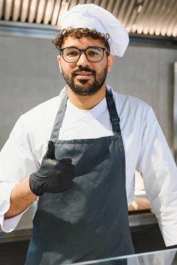 Genç aşçı siyah eldiven ve önlük takıyor. Profesyonel bir mutfakta çalışırken baş parmak işareti yapıyor. Doğu mutfağında uzmanlaşıyor.